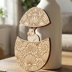 Peut inclure: Décoration en bois en forme d'œuf de Pâques avec un lapin à l'intérieur. L'œuf est divisé en deux moitiés, avec des motifs de coquillages et géométriques. Le lapin est brun clair, et l'œuf est posé contre un vase blanc.