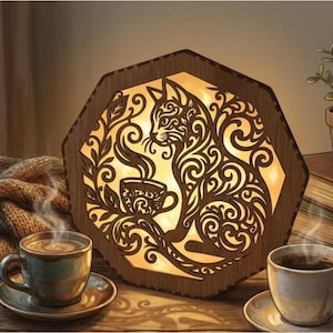 Könnte beinhalten: Eine hölzerne Katzenlampe mit warmem Schein, die eine Katze und eine Kaffeetasse zeigt. Zwei Kaffeetassen und eine kleine Topfpflanze sind ebenfalls zu sehen. Die Lampe steht auf einer Holzoberfläche und schafft eine gemütliche Atmosphäre.