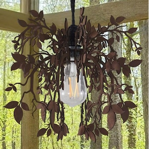 Puede incluir: Lámpara colgante decorativa con diseño de hojas de metal marrón. La lámpara rodea una bombilla de vidrio transparente con filamento visible. La luz cuelga de un cable negro, creando una estética inspirada en la naturaleza.