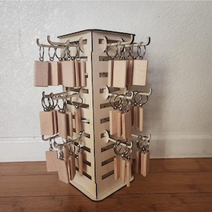 May include: A wooden rotating display stand with multiple hooks holding rectangular wooden keychains with metal rings. The stand is made of light-colored wood and is designed to showcase keychains or similar small items.