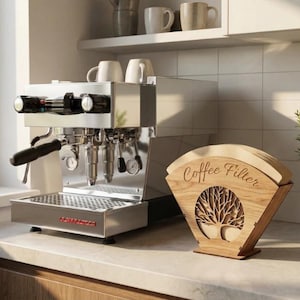 May include: A stainless steel espresso machine with black accents sits on a countertop next to a wooden coffee filter holder. The holder has a tree design and the words "Coffee Filter" are engraved on it.