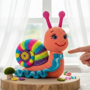 May include: A handmade crochet snail toy with a smiling face, pink antennae, and a colorful shell. The snail's body is peach-colored, with a blue scarf and a ruffled blue base. It sits on a wooden slice with decorative elements.