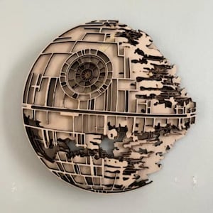 May include: A wooden Death Star wall art piece. The artwork is a layered design with a detailed circular center and a partially destroyed outer surface. The color is a natural wood tone with darker edges, creating a 3D effect.