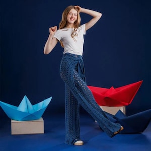 May include: A woman in a white t-shirt and blue crochet trousers. The trousers have a wide leg and a tie waist. The background is a dark blue with paper boat decorations in blue and red.