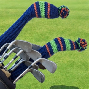 Könnte beinhalten: Eine Golftasche mit mehreren Schlägern, teilweise sichtbar, und zwei blauen Häkel-Schlägerhauben mit grünen und orangefarbenen Streifen und einem Bommel. Der Hintergrund ist ein grünes Feld.