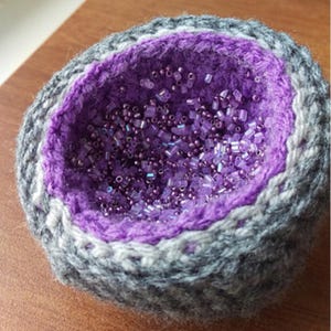 Op de afbeelding: Een handgemaakte, gehaakte kom met een geode-achtig ontwerp. De buitenkant is grijs en wit, met een levendig paars interieur gevuld met kleine, iriserende kralen. De textuur en het kleurenpalet van de kom creëren een uniek decoratief stuk.