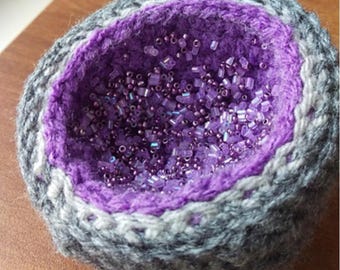 May include: A handmade, crocheted bowl with a geode-like design. The exterior is gray and white, framing a vibrant purple interior filled with small, iridescent beads. The bowl's texture and color palette create a unique decorative piece.