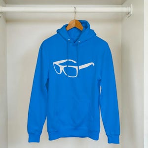 Può includere: Una felpa con cappuccio blu brillante con una grafica di occhiali bianchi. La felpa ha un cappuccio con coulisse e una tasca frontale. È appesa a un'appendiabiti di legno all'interno di un armadio bianco.