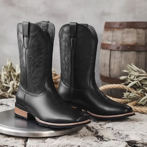 Puede incluir: Un par de botas de vaquero negras con intrincados diseños cosidos en la caña. Las botas tienen una puntera cuadrada, un tacón bajo y una suela marrón claro. Se exhiben sobre una superficie metálica, con un barril rústico y una cuerda en el fondo.