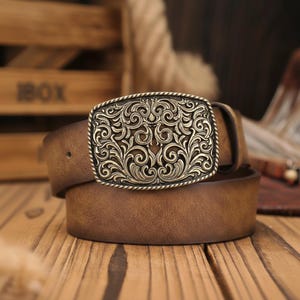 Puede incluir: Un cinturón de cuero marrón con una hebilla rectangular decorativa de oro antiguo. La hebilla presenta un diseño floral ornamental y arremolinado. El cinturón está enrollado sobre una superficie de madera.