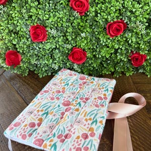 May include: A floral-patterned book cover with a light pink ribbon. The cover features a white background with pink flowers, green leaves, and yellow accents. Red roses are in the background.