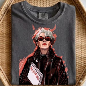 Pode incluir: T-shirt cinzento escuro com uma ilustração estilizada de uma mulher usando óculos de sol e uma jaqueta preta, segurando um livro intitulado "RUNWAY". O design inclui detalhes em vermelho e uma gola de pele sintética. A camiseta é da Comfort Colors.
