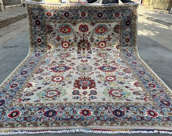 May include: A large rectangular area rug featuring a light gray field with a symmetrical floral pattern in shades of red, blue, green, and cream. The rug has a wide blue border with intricate floral motifs and white fringe at the ends.