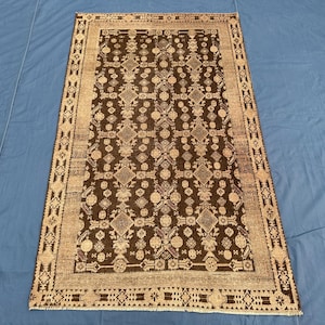 May include: A rectangular, brown area rug with a detailed geometric pattern. The rug features a central field of dark brown with repeating diamond and floral motifs. A lighter tan border frames the design, adding contrast. The rug is placed on a blue surface.