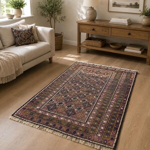 May include: A rectangular area rug featuring a central field of muted brown and mauve tones with a repeating geometric pattern of diamonds and crosses. The rug is framed by a multi-layered border in shades of blue, brown, and cream with fringe.