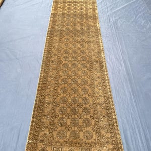 May include: A long, narrow runner rug featuring a repeating geometric pattern in shades of tan, beige, and dark brown. The rug has a central field filled with octagonal motifs and a decorative border with intricate linear designs and fringe ends.