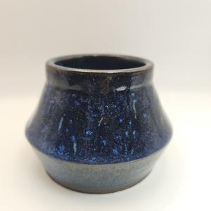 May include: A small, handcrafted ceramic vase with a dark blue, speckled glaze. The vase has a wide, rounded body that tapers slightly towards the base, with a dark rim. The lower portion of the vase is a muted gray-blue.