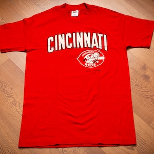 May include: Red t-shirt with a white "Cincinnati Reds" logo featuring a cartoon mascot. The logo includes the words "Cincinnati Reds" and a cartoon mascot.
