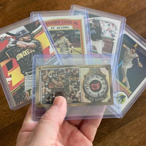 May include: A collection of baseball trading cards featuring Fernando Tatis Jr. and other players. The cards are in protective sleeves, showcasing action shots and team logos. The cards are in various designs and colors.