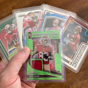 May include: A hand holding several football trading cards in protective sleeves. The cards feature players in red and gold uniforms. One card is labeled "Premier" with the name "Jacob Cowing". The cards are in clear plastic holders.