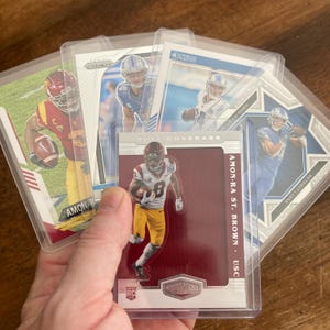 May include: A collection of football trading cards featuring players in action. The cards are encased in protective sleeves. One card prominently displays "AMON-RA ST. BROWN" and "USC" with a player in a USC uniform. Other cards show players in Detroit Lions uniforms.