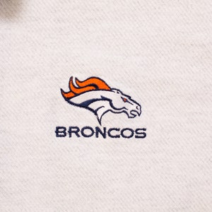 May include: White polo shirt with a navy blue embroidered Denver Broncos logo. The logo features a white horse with an orange flame mane and the word "BRONCOS" in navy blue.