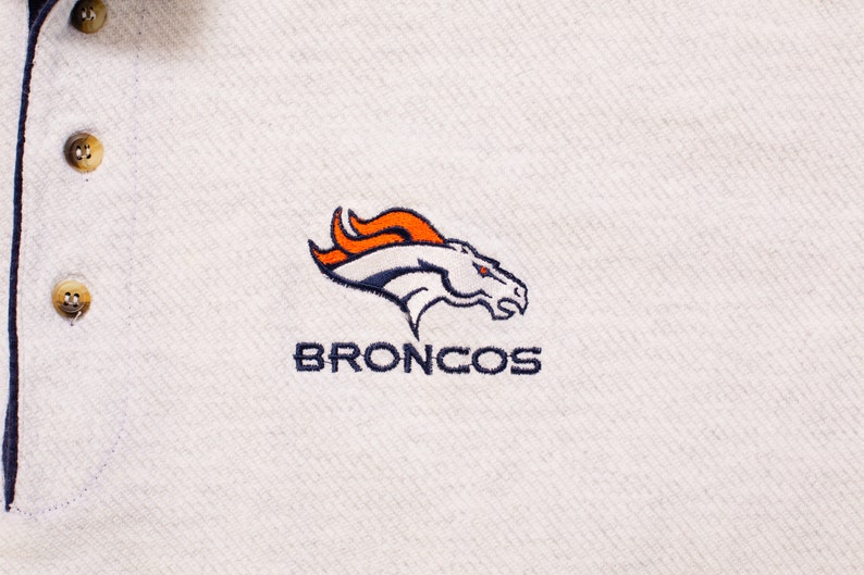 May include: White polo shirt with a navy blue embroidered Denver Broncos logo. The logo features a white horse with an orange flame mane and the word "BRONCOS" in navy blue.