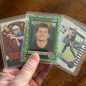 May include: Three football trading cards in protective sleeves. One card features a player with a guitar graphic, another has a player with the text "RISING STARS", and the third shows a player throwing a football. The cards are in clear plastic holders.