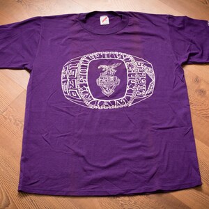 May include: Purple t-shirt with a white graphic of a championship ring with the words "1980 NCAA CHAMPIONS FIGHTERS" around the ring.