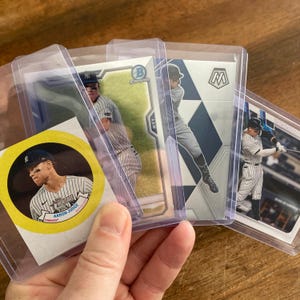 May include: A collection of baseball trading cards featuring New York Yankees players, including Aaron Judge. The cards are in protective sleeves, showcasing various designs and brands. The cards are held in a hand.