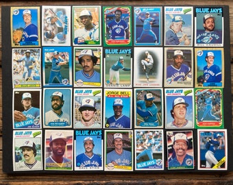 May include: A collection of 24 baseball trading cards featuring Toronto Blue Jays players. The cards are arranged in a grid on a dark brown surface. Each card features a player's photo, name, and team logo. The cards are in good condition and have a vintage feel.