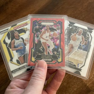 May include: Three basketball trading cards featuring Angel Reese. The cards are in protective sleeves. One card has the text "MONOPOLY" and "SKYTO S". The center card has a red background and the text "ANGEL REESE". The third card says "ALL-AMERICAN".