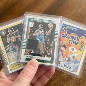 May include: Three basketball trading cards featuring Angel Reese of the Chicago Sky. The cards are in protective sleeves. One card is from the "Jersey Series" and another features "Crunch Time" text. The cards showcase action shots of Reese.