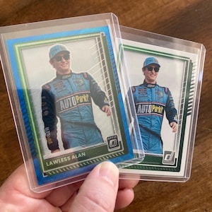May include: Two trading cards featuring Lawless Alan in a blue racing suit, held in protective sleeves. The cards have a blue and green border, with the "Optic" and "Donruss" logos visible. The text "AUTO Parkit" is on the suit.