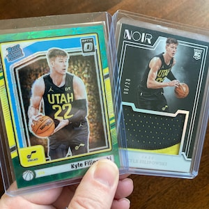 May include: Two basketball trading cards in protective sleeves. One card is green with the text "RATED ROOKIE" and "OPTIC" and features a player in a Utah Jazz uniform. The other card is black with the text "NOIR" and features the same player.