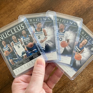 May include: A collection of basketball trading cards featuring Minnesota Timberwolves players. The cards are in protective sleeves and display the "Origins" and "Nucleus" branding. Players are shown in action with basketballs.