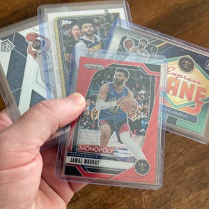 May include: A collection of basketball trading cards featuring Jamal Murray and other players. The cards are in protective sleeves. One card has the text "MONOPOLY" and "JAMAL MURRAY". Another card has the text "Express LANE".
