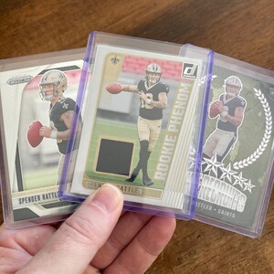 May include: Three football trading cards featuring Spencer Rattler in a New Orleans Saints uniform. The cards are in protective sleeves. One card is labeled "ROOKIE PHENOM". The cards are in shades of black, gold, white, and green.