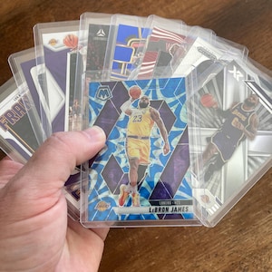 Puede incluir: Una colección de cromos de baloncesto en fundas protectoras. Las cartas muestran a jugadores en acción, incluido LeBron James con el uniforme de los Lakers. Las cartas tienen varios diseños y colores, siendo el morado, el dorado y el azul los colores dominantes.
