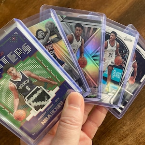 May include: A collection of five basketball trading cards featuring Brooklyn Nets players. The cards are in protective plastic sleeves. The cards feature different players, including Cameron Thomas, who is wearing a jersey with the number 8. The cards have a variety of designs, including a green and blue knit pattern, a holographic design, and a white and blue design.
