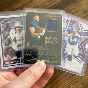 May include: Three sports trading cards featuring Cam Skattebo. The left card shows a player in a white jersey with the number 4. The center card is a black Rookies & Stars card with a blue fabric swatch and a crown graphic. The right card shows a player in a blue jersey with the number 44.