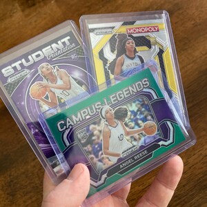 May include: Three basketball trading cards in protective plastic cases. The top card features a female basketball player in a blue and white uniform with the text "PRIZM MONOPOLY" on the card. The middle card features a female basketball player in a purple and white uniform with the text "STUDENT ORIENTATION" on the card. The bottom card features a female basketball player in a white and purple uniform with the text "CAMPUS LEGENDS ANGEL REESE" on the card.