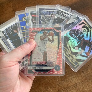 May include: A hand holding a collection of basketball trading cards in protective sleeves. The cards feature images of players in action, with team logos and player names visible. The cards are in clear plastic holders.