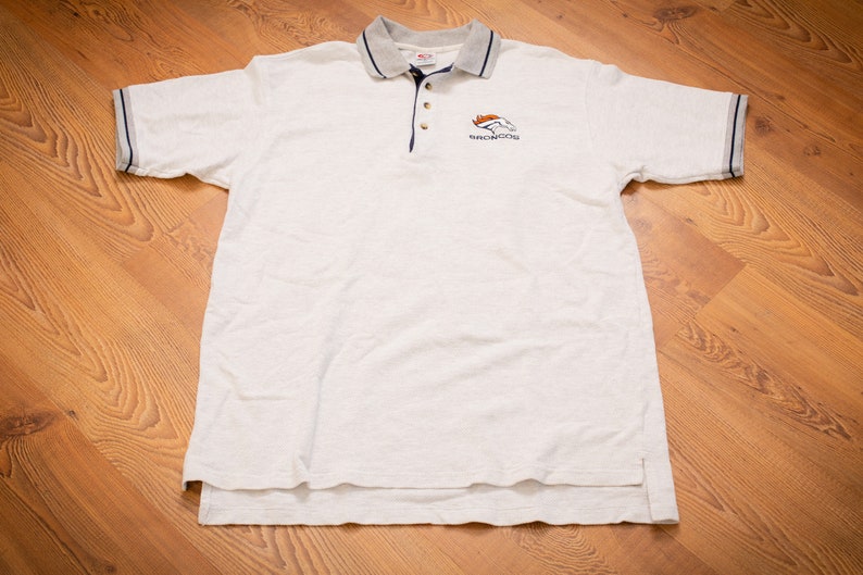 May include: White polo shirt with a navy blue trim and a small embroidered Denver Broncos logo on the chest.