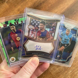 May include: Three trading cards featuring Ronald Araujo. The cards are in protective sleeves. One card has a green background with the Spotify logo. Another card has a FIFA logo and the player's signature. The third card has a blue background.