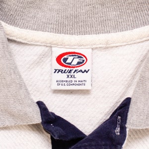May include: White polo shirt with a gray collar and navy blue trim. The shirt has a red and white label that says "TRUEFAN" and "XXL" with the text "Assembled in Haiti of U.S. Components".