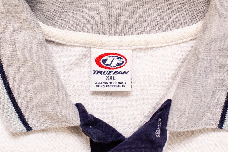 May include: White polo shirt with a gray collar and navy blue trim. The shirt has a red and white label that says "TRUEFAN" and "XXL" with the text "Assembled in Haiti of U.S. Components".