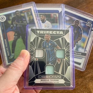 May include: A collection of Marcus Thuram trading cards in protective sleeves. The cards feature images of the soccer player in his FC Internazionale Milano uniform. One card is labeled "TRIFECTA" and "OBSIDIAN".