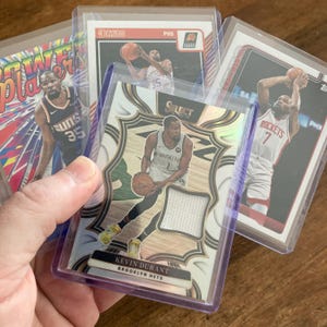 May include: A collection of basketball trading cards featuring Kevin Durant in various team uniforms. The cards are encased in protective sleeves. One card has a jersey swatch. The cards showcase Durant playing for the Suns, Nets, and Rockets.