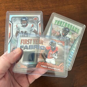 May include: A collection of football trading cards in protective cases. One card features the text "FIRST YEAR FABRIC" and a jersey swatch. Other cards display players in action, with team colors and logos. The cards are held together.